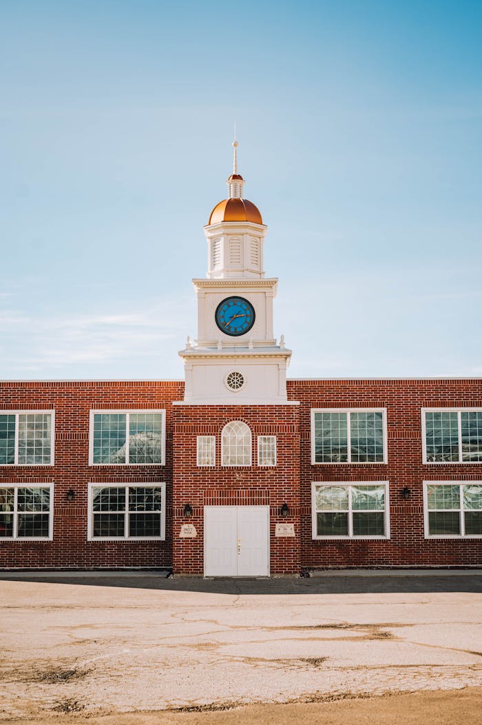 contact-img Sunlit clock tower on a vintage brick building under a clear blue sky.